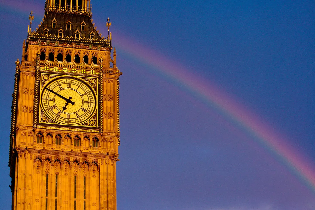 The Elizabeth Tower clock face lit gold by the evening sun with a rainbow behind