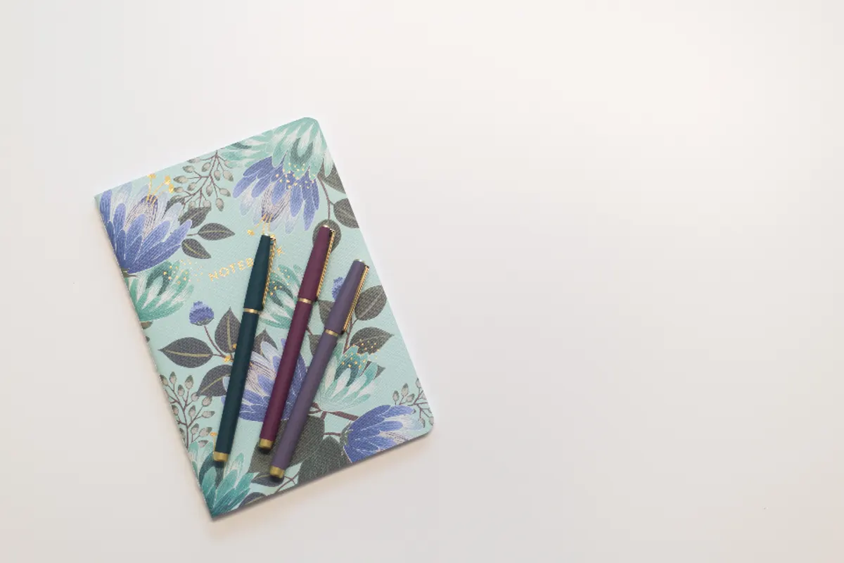 A floral-covered notebook with three coloured pens lying on top, on a white desk