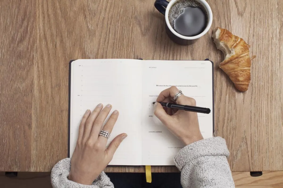 Hands writing a list in an open notebook beside a cup of black coffee and a croissant