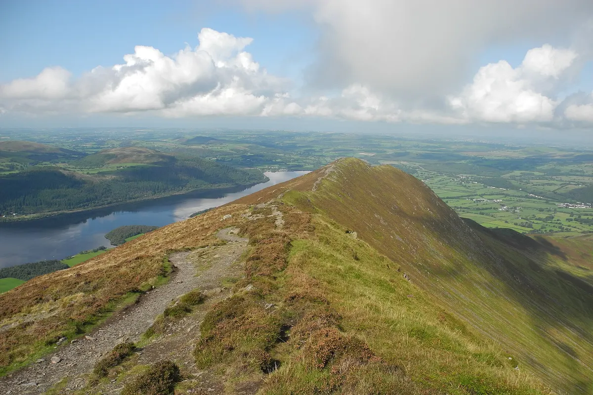 A grassy ridge path in the Lake District leading towards a lake and green valley under summer clouds