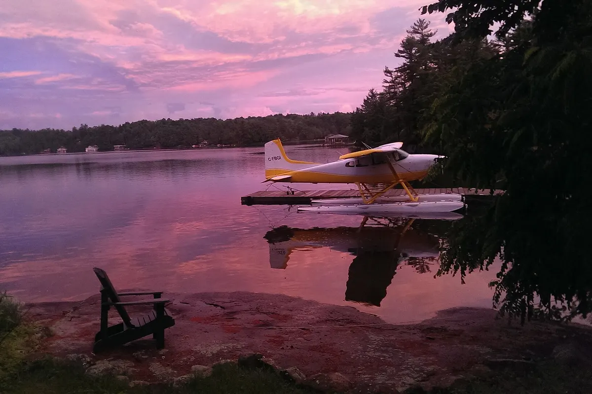 A floatplane moored at a cottage dock on a lake at pink dusk, with a wooden chair on the shore