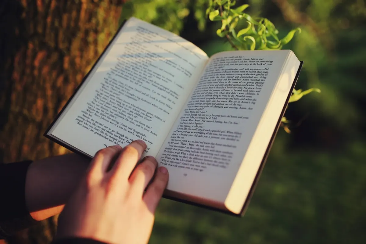 A hand holding an open book outdoors with green leaves behind it