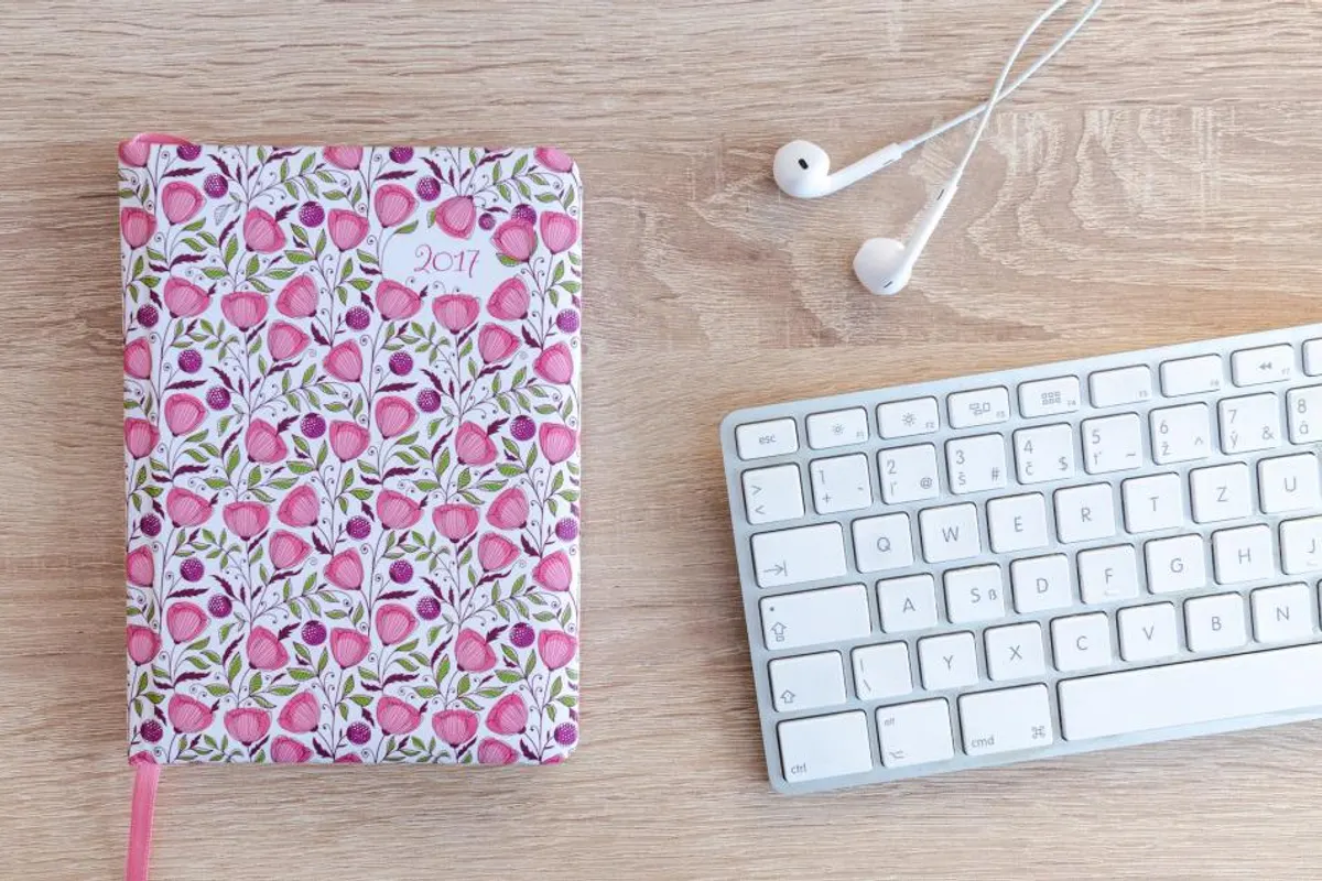 A pink floral notebook beside a white keyboard and earphones on a wooden desk