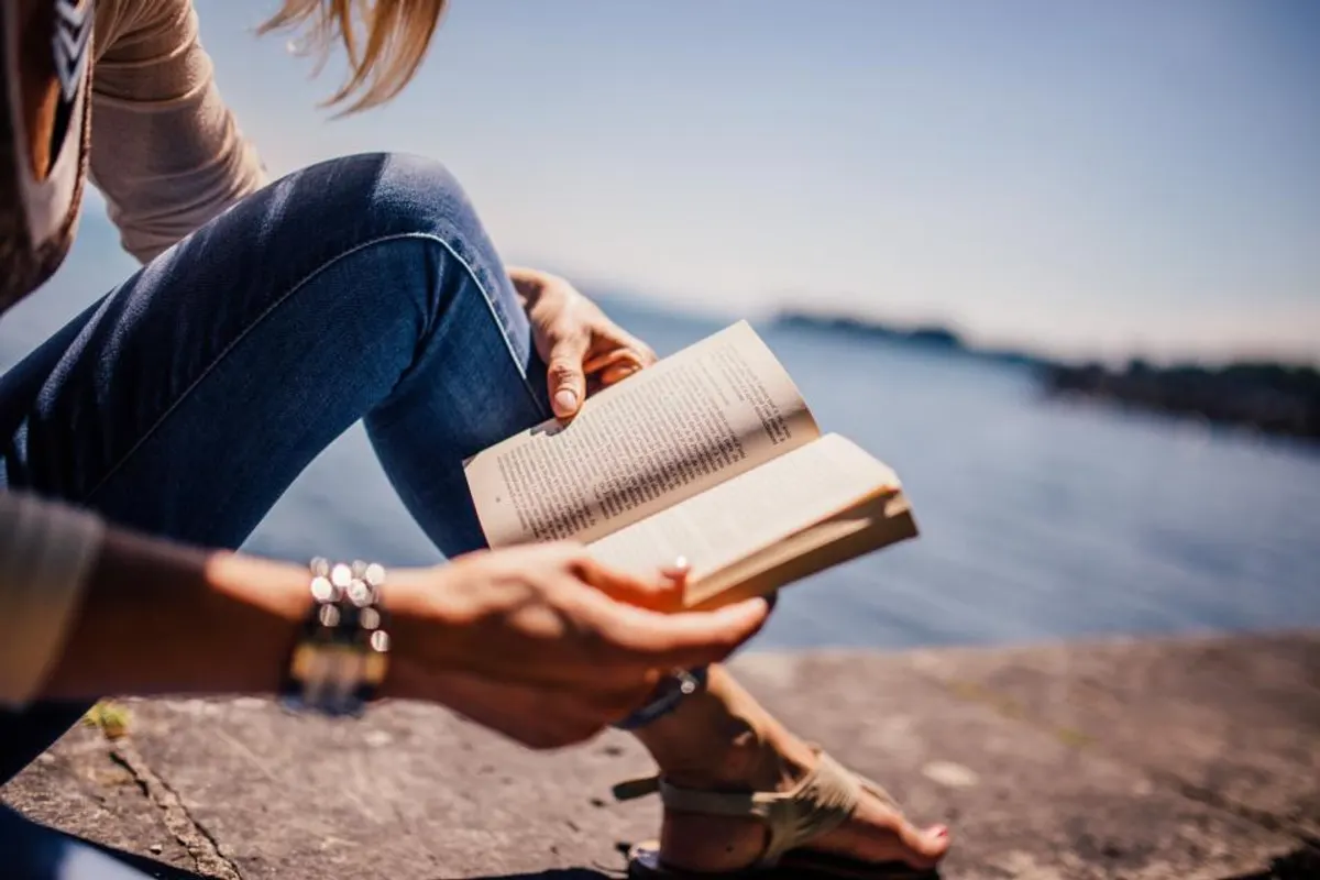 Someone sitting by a lake holding an open paperback book