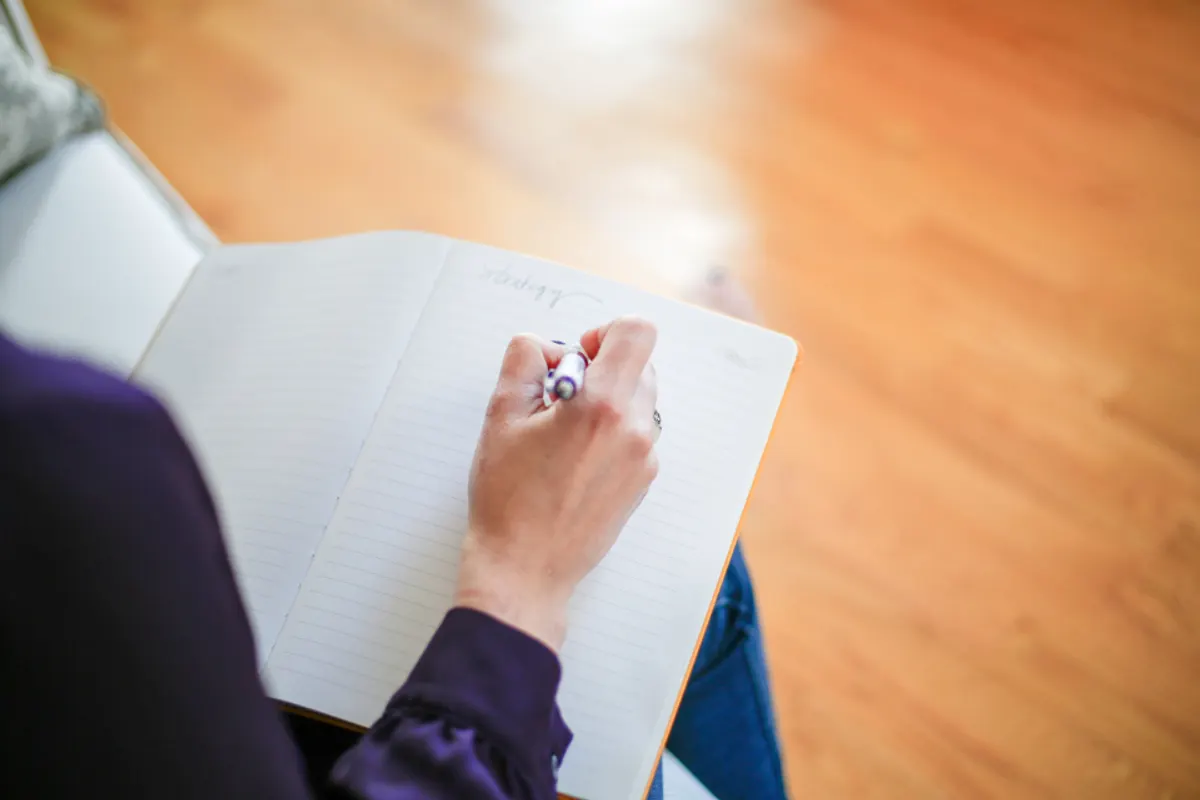 A hand writing in a notebook resting on someone’s lap, in warm light